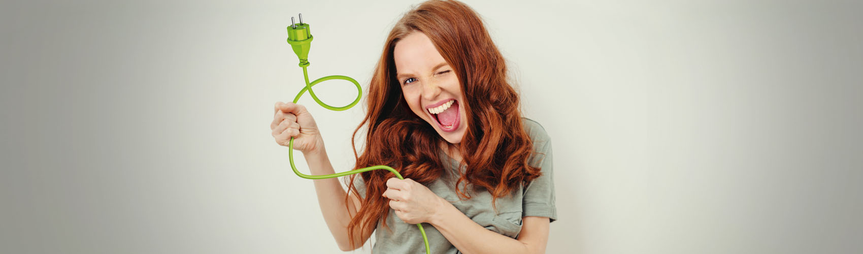 Frau lächelt mit einem zugekniffenen Auge und aufgerissenem Mund in die Kamera und hält ein grünes Stromkabel